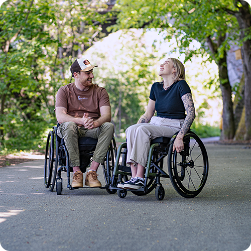 A young woman and a young man sit in wheelchairs and laugh at each other.